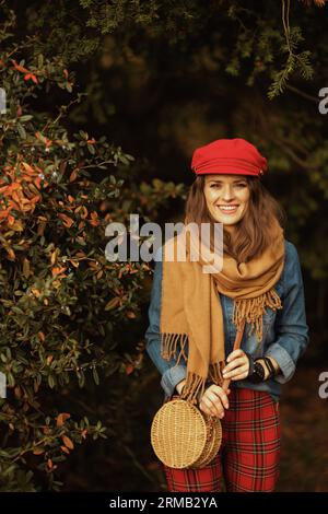 Hallo Herbst. Lächelnde moderne Frau in Jeanshemd und roter Mütze mit Schal, Handschuhen und Tasche im Stadtpark. Stockfoto