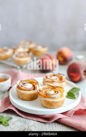 Nektarinenrosen aus Blätterteig, auf einer weißen Platte. Buttrige, flakige und knusprige Blätterteigrollen mit Schichten aromatischer Nektarinscheiben. Pinkfarbenes Nickerchen Stockfoto