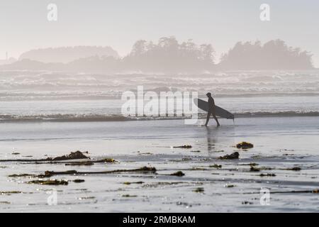 Unerkennbare Surfer-Silhouette mit Surfbrettwanderungen am Cox Bay Beach am Pazifik, Tofino, Vancouver Island, BC, Kanada. Stockfoto