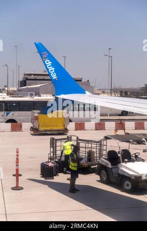 Flughafenarbeiter laden Gepäck von FlyDubai aus in ein Gepäckfahrzeug. Flughafen Dubai, VAE. Flughafen Cargo Loading Cars, Gepäcktraktor-Anhänger Stockfoto