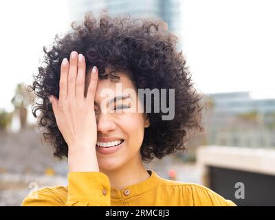 Junge, unbekümmerte, afroamerikanische lächelnde Frau, die in die Kamera schaut, während sie mit der Hand ein Auge bedeckt. Stockfoto
