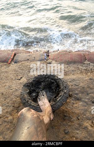 Der Fuß des Mannes in der Schlammschale, das Bein des Mannes bedeckt mit Schlamm. Schüssel mit Schlamm zum Auftragen auf den Körper vor dem Schwimmen im Toten Meer Jordan am Strand Stockfoto