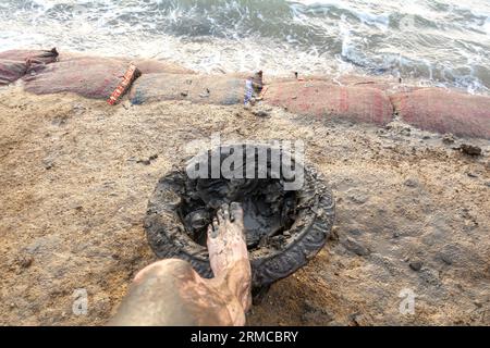 Der Fuß des Mannes in der Schlammschale, das Bein des Mannes bedeckt mit Schlamm. Schüssel mit Schlamm zum Auftragen auf den Körper vor dem Schwimmen im Toten Meer Jordan am Strand Stockfoto