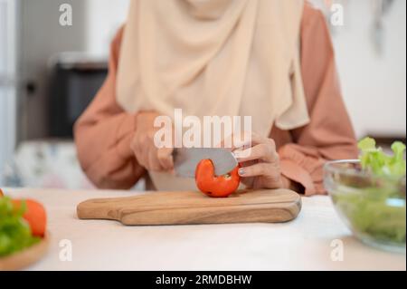 Nahaufnahme einer muslimischen Frau, die Zutaten für ihre Salatmischung in einer Küche zubereitet, eine Tomate auf einem Schneidebrett schneidet und in der Küche kocht Stockfoto