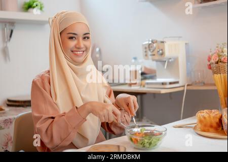 Eine schöne und lächelnde asiatische Muslimin oder Hausfrau in einem Hijab mischt einen gesunden Salat in einer Schüssel, macht ihre gesunde Salatschale, und isst es ich Stockfoto