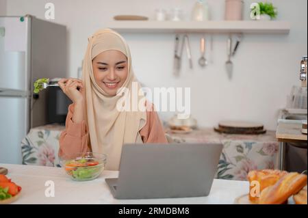 Eine schöne, glückliche asiatische Muslimin isst ihren gesunden Salat, während sie an ihrem Laptop an einem Esstisch in der Küche arbeitet. Arbeiten Sie von zu Hause aus Stockfoto
