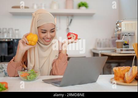 Eine glückliche und schöne asiatische Muslimin in einem Hijab arbeitet an ihrem Laptop an einem Esstisch in der Küche. Arbeiten Sie von zu Hause aus, Food Blogger, Inland Stockfoto