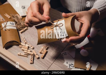 Eine nicht wiedererkennbare junge Frau klebt die Nummer auf die Handwerkstasche, wird mit einer Wäscheklammer befestigt. Weiblich macht Kraftpapier für hausgemachte Adventskalender mit eigenen Händen Schritt für Schritt selbst gemacht. Vorbereitung auf das weihnachtskonzept. Saisonale Aktivitäten für Kinder, Familienurlaub im Winter. Umweltfreundlich Geschenke. Öffnen Sie das Paket jeden Tag Stockfoto