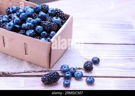 Heidelbeeren und Brombeeren in einer Pappschachtel auf einem Holztisch. Mischung aus blauen Beeren aus nächster Nähe. Stockfoto