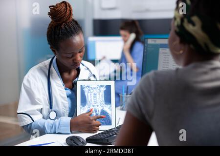Radiologe zeigt Röntgen-ct-Lungen-Scan und erklärt dem Patienten während des Termins klinische Informationen auf der Tablette. Arzt im Krankenhaus mit Radiologie und afroamerikanische Frau bei ärztlicher Untersuchung. Stockfoto