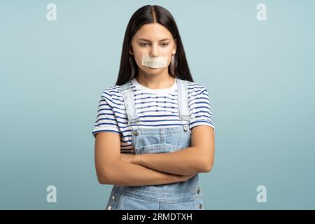 Trauriges frustriertes Teenager-Mädchen mit geschlossenem Bandmund auf türkisfarbenem Hintergrund, der nach unten schaut. Stockfoto