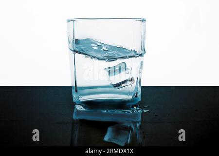 Ein Eiswürfel fällt in ein Glas Wasser. Schwarz-weißer Hintergrund. Turbulentes Wasser im Schiff. Stockfoto