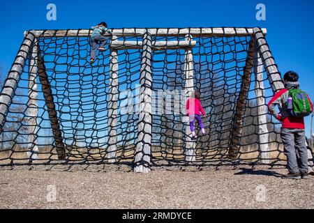 Zwei kleine Kinder klettern draußen an die hohe Seilwand, während Vater anschaut Stockfoto