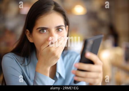 Eine schockierte Frau, die ein Smartphone in einer Bar hält, sieht dich an Stockfoto