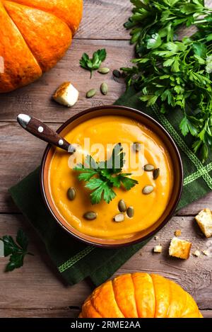 Kürbissuppe mit Petersilie, Kürbiskernen und Croutons und Bio-Kürbissen auf hölzernem Hintergrund, Draufsicht. Saisonale Herbstmahlzeit, Komfortessen. Stockfoto