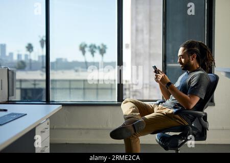 Seitenansicht des Geschäftsmannes, der mit dem Smartphone unterwegs ist, während er auf dem Stuhl gegen das Fenster im Büro sitzt Stockfoto
