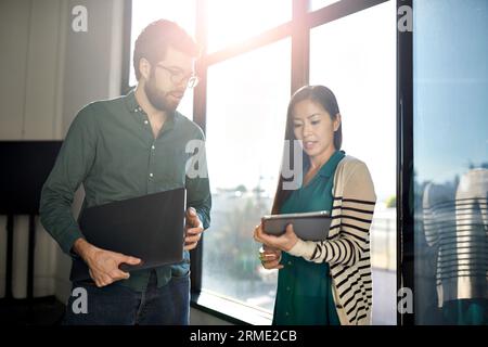Geschäftsfrau zeigt dem Geschäftsmann einen Tablet-Computer, während er im Büro vor dem Fenster steht Stockfoto