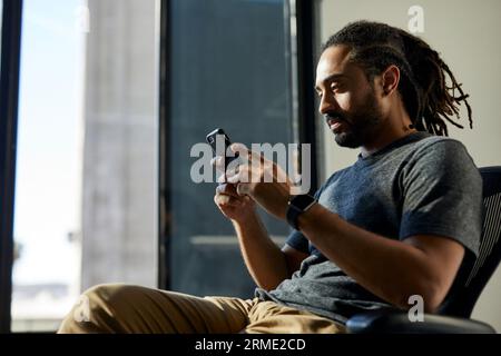 Seitenansicht eines Geschäftsmannes mit Dreadlocks über das Smartphone, während er auf dem Stuhl gegen das Fenster im Büro sitzt Stockfoto