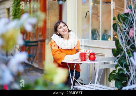 Glückliche junge Frau in Paris an einem Wintertag, trinken Kaffee, Tee oder heiße Schokolade im Pariser Café im Freien, das zu Weihnachten dekoriert ist, und schreiben Postkarten Stockfoto