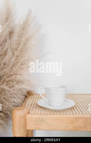 Eine weiße Kaffeetasse auf einer Untertasse auf einer Rattanbank mit Pampas gra Stockfoto