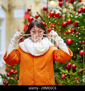 Glücklicher junger Tourist in Paris an einem Wintertag, der Spaß mit weißen und roten Zuckerstangen hat, geschmückter Weihnachtsbaum ist im Hintergrund Stockfoto