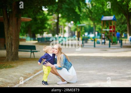 Die schöne junge Mutter und ihr entzückender Kleinkind-Sohn haben Spaß zusammen auf einem Pariser Spielplatz, Junge küsst seine Mami Stockfoto