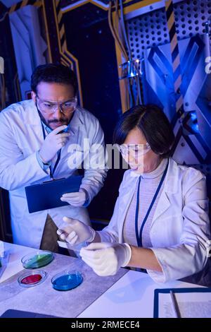 indischer Wissenschaftler mit Klemmbrett, der Laborglas in der Nähe von Kollegen und Petrischalen betrachtet Stockfoto