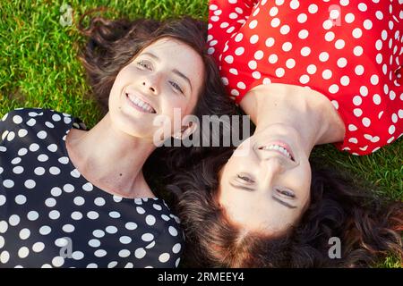 Wunderschöne Zwillingsschwestern in roten und schwarzen Polka-Punkt-Kleidern, die auf dem Gras nahe beieinander liegen und lächeln Stockfoto