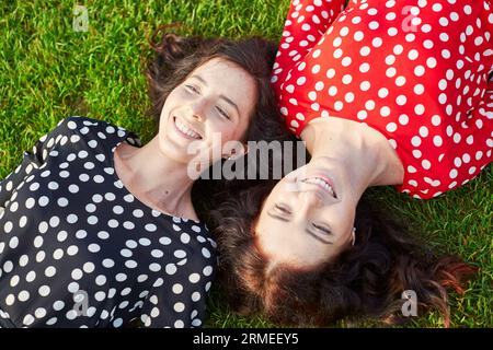 Wunderschöne Zwillingsschwestern in roten und schwarzen Polka-Punkt-Kleidern, die auf dem Gras nahe beieinander liegen und lächeln Stockfoto