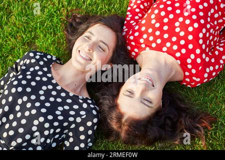 Wunderschöne Zwillingsschwestern in roten und schwarzen Polka-Punkt-Kleidern, die auf dem Gras nahe beieinander liegen und lächeln Stockfoto