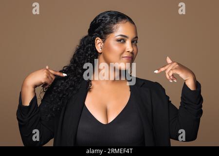 Das bin ich. Selbstbewusste, stolze brasilianische und große Frau, die mit den Fingern auf braunen Hintergrund zeigt Stockfoto