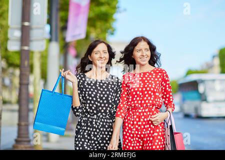 Glückliche Zwillingsschwestern, die während ihres Urlaubs in Frankreich shoppen gehen, mit Einkaufstaschen vor dem Arc de Triomphe auf der Champs-Elysees, Paris, Frankreich Stockfoto