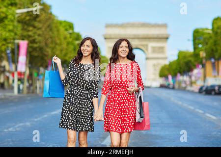 Glückliche Zwillingsschwestern, die während ihres Urlaubs in Frankreich shoppen gehen, mit Einkaufstaschen vor dem Arc de Triomphe auf der Champs-Elysees, Paris, Frankreich Stockfoto