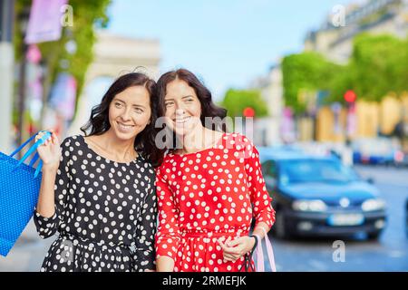 Glückliche Zwillingsschwestern, die während ihres Urlaubs in Frankreich shoppen gehen, mit Einkaufstaschen vor dem Arc de Triomphe auf der Champs-Elysees, Paris, Frankreich Stockfoto