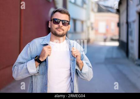 Gutaussehender Mann mit Sonnenbrille und Jeansjacke, der vor der Straße der europäischen Stadt posiert. Stockfoto