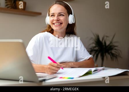 Junge Schülerin, die zu Hause Hausaufgaben macht, hört pädagogische Podcasts mit ihren Kopfhörern und ihrem Laptop. Stockfoto