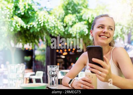 Sommer-City-Lifestyle, glückliche Frau, die im Straßencafé sitzt, ihr Telefon benutzt und lächelt. Stockfoto