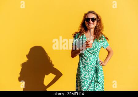 Junge lockige Frau, die ein trendiges grünes Kleid und eine Sonnenbrille trägt, die ein Glas Saft in der Hand hält und in der Nähe eines gelben Hintergrunds posiert. Stockfoto
