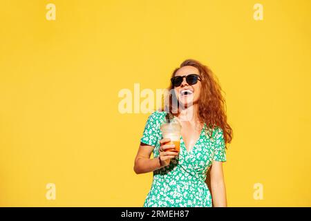 Fröhliche junge Lockenhaarfrau in der Sonnenbrille, die ein Glas Saft in der Hand hält und lachend. Stockfoto