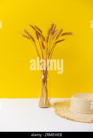 Stillebenstrauß aus gelbem Weizen in Vase aus Glasflasche und Strohsonnenhut auf weißem Tisch vor gelbem Hintergrund. Stockfoto