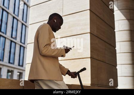 Seitenansicht eines jungen afroamerikanischen Geschäftsmannes mit Kopfhörern und Anzug, der sich im Mobiltelefon die Playlist anschaut, während er am Gebäude steht Stockfoto