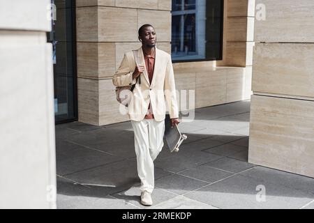 Junger, seriöser Geschäftsmann in Pastellfarben, der Skateboard trägt, während er sich am Morgen entlang der Säulen eines modernen Gebäudes bewegt Stockfoto