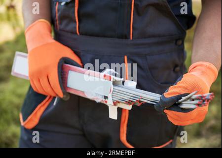 Nahaufnahme von Handschuhen in orangefarbenen Arbeitshandschuhen, die Schweißelektroden für die Installation von Solarpaneelen halten. Männlicher Baumeister mit beschichteten Metalldrähten oder Schweißstäben in den Händen. Stockfoto