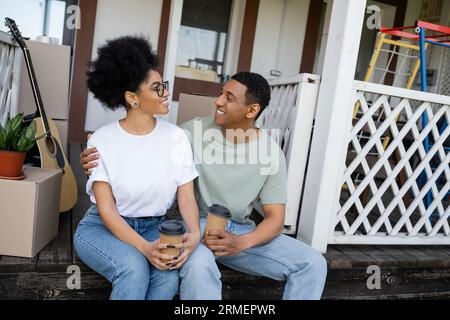 Positives afroamerikanisches Paar umarmt und hält Kaffee, um in die Nähe von Boxen auf der Veranda des neuen Hauses zu gehen Stockfoto