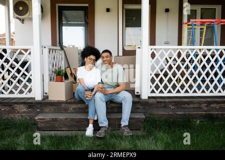 Fröhliches afroamerikanisches Paar, das sich umarmt und Kaffee hält, um auf die Veranda des neuen Hauses zu gehen Stockfoto