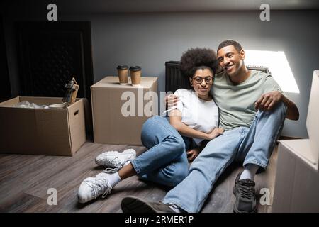 Lächelndes romantisches afroamerikanisches Paar, das sich in der Nähe von Kaffee und Kisten auf dem Dachboden in einem neuen Haus umarmt Stockfoto