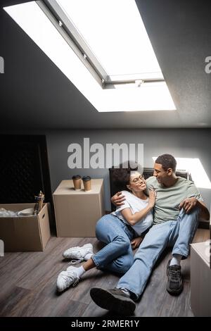 Lächelndes afroamerikanisches Paar umarmt sich in der Nähe von Kaffee und Pappkartons auf dem Dachboden in einem neuen Haus Stockfoto
