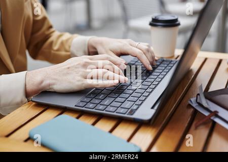 Hände einer jungen Geschäftsfrau oder eines Solopreneurs, die auf der Laptop-Tastatur tippen, während sie am Holztisch im Café im Freien sitzen und Arbeit organisieren Stockfoto