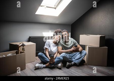 Lächelndes afroamerikanisches Paar, das Kaffee hält, während es in der Nähe von Kästen auf dem Dachboden in einem neuen Haus sitzt Stockfoto