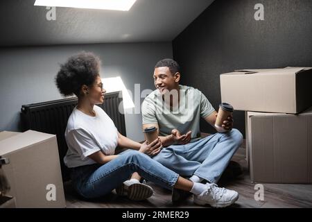 Fröhliches afroamerikanisches Paar mit Kaffee, der in der Nähe von Pappkartons auf dem Dachboden in einem neuen Haus spricht Stockfoto